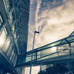 Moderner gläserner Wolkenkratzer und erhöhter Gehweg mit einer Straßenlaterne im Vordergrund vor einem dramatischen Himmel mit Sonnenlicht und sichtbaren Kondensstreifen von Flugzeugen.