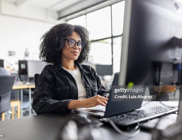 Eine Frau mit lockigem Haar und Brille sitzt an einem Schreibtisch in einem Büro und arbeitet an einem Computer. Sie ist auf ihren Bildschirm konzentriert, während im Hintergrund natürliches Licht aus großen Fenstern fällt.