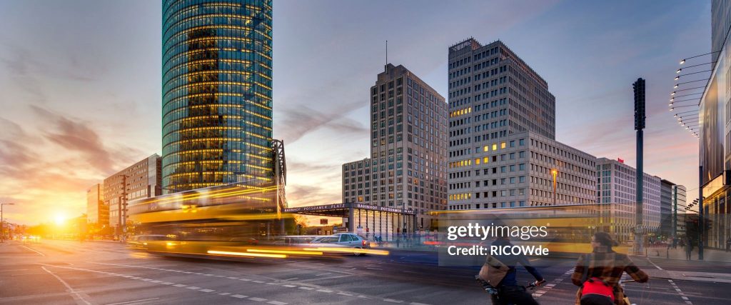 Belebte Stadtkreuzung bei Sonnenuntergang mit hohen modernen und klassischen Gebäuden, einem vorbeifahrenden gelben Bus, Radfahrern im Vordergrund und einem strahlenden Himmel im Hintergrund.