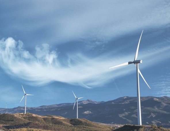 Mehrere große weiße Windräder stehen auf einem grasbewachsenen Hügel mit Bergen im Hintergrund unter einem blauen Himmel mit Wolkenfetzen.