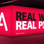 Eine Person geht an einer großen, hell erleuchteten rot-weißen Anzeige vorbei, die ein fettes A-Logo, einen Teil des Textes mit der Aufschrift REAL WOR und REAL PEOP sowie die Website Adecco.com zeigt.