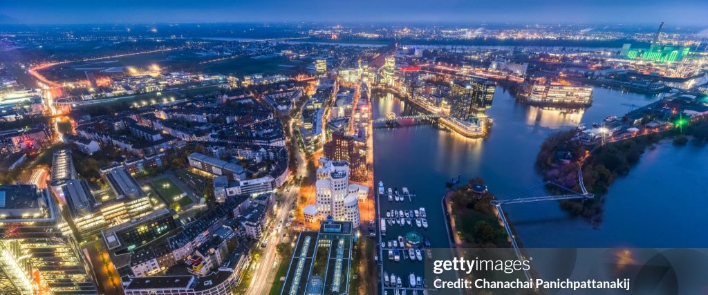 Nachtansicht einer Stadt aus der Vogelperspektive mit beleuchteten Gebäuden entlang eines Flusses, angedockten Booten und weit entfernten glühenden Lichtern unter einem blauen Dämmerungshimmel.
